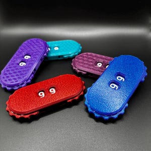 Puede incluir: Una colección de objetos ovalados y texturizados de colores, cada uno con el número "99" en blanco. Los objetos son en tonos de rojo, morado, azul, turquesa y morado oscuro. Los objetos tienen un borde acanalado.