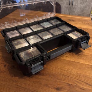 Könnte beinhalten: Eine schwarze Kunststoff-Kartenaufbewahrungsbox mit transparentem Deckel, gefüllt mit Sammelkarten. Die Box hat mehrere Fächer und einen grauen Griff. Die Karten sind durch den transparenten Kunststoff sichtbar, auf einer Holzoberfläche.