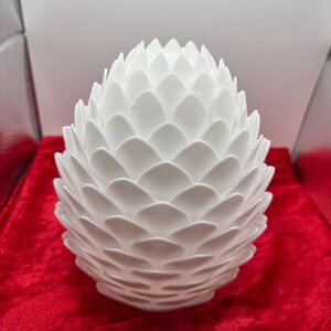 May include: A white ceramic dragon egg with a textured, scaly surface. The egg is shaped like a traditional dragon egg with a pointed top and a rounded bottom.