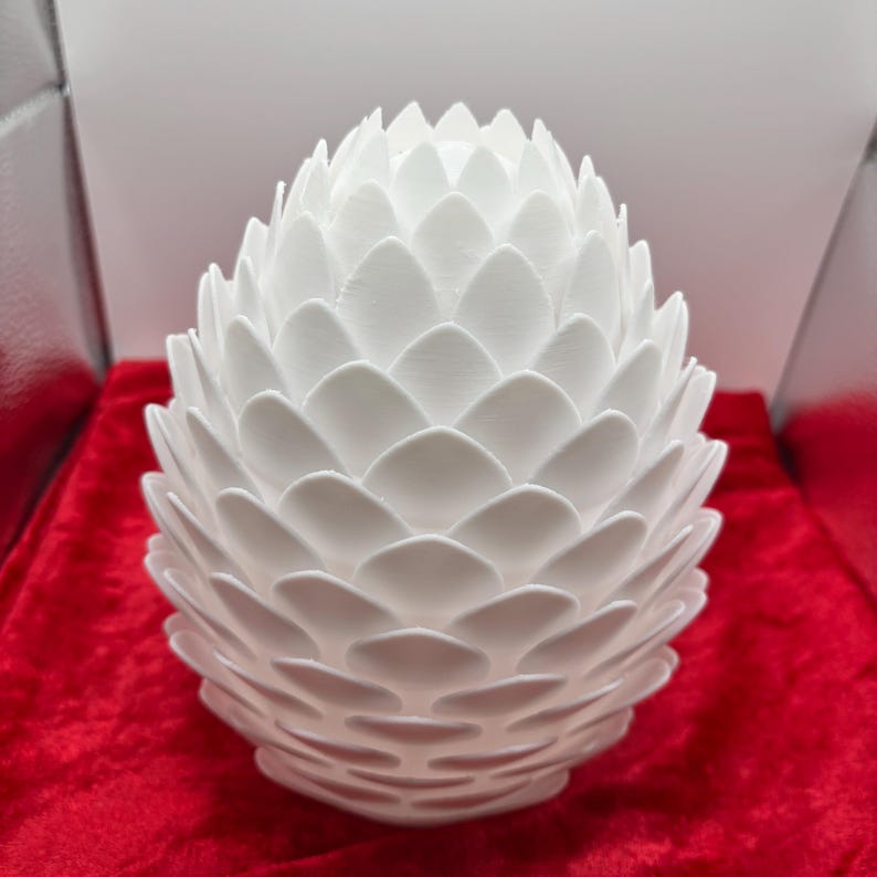 May include: A white ceramic dragon egg with a textured, scaly surface. The egg is shaped like a traditional dragon egg with a pointed top and a rounded bottom.