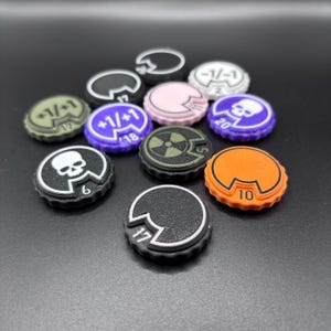 以下が含まれることがあります： 黒、紫、オレンジ、オリーブグリーン、ピンクなど、様々な色の円形ゲームトークンのコレクション。各トークンには刻み目のある縁があり、ドクロ、放射能マーク、または+1/+1、-1/-1、6、10、17、18、20といった数字などの中心グラフィックが描かれています。