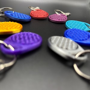 Puede incluir: Una colección de llaveros coloridos en varios tonos, incluyendo azul, rojo, morado, amarillo y turquesa. Cada llavero presenta una etiqueta texturizada en forma ovalada unida a un llavero plateado. Los llaveros están sobre una superficie oscura.