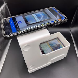 Könnte beinhalten: Ein weißes Kunststoff-Kartenscannergerät mit einem Smartphone obenauf. Das Telefon zeigt ein Kartenbild an. Eine Box darunter enthält mehrere Sammelkarten. Das Gerät dient zum Scannen und Organisieren von Sammelkarten.