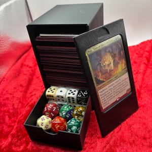 Peut inclure: Boîte de rangement de cartes noire avec un compartiment pour dés. La boîte contient un jeu de cartes et une variété de dés de différentes couleurs. Une carte avec le texte "Voja, Jaws of the Conclave" est visible.