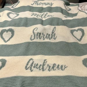 May include: A handmade, crocheted blanket in cream and teal. The blanket features horizontal stripes with names and heart shapes. The names "Thomas", "Miller", "Sarah", and "Andrew" are displayed in teal script across the blanket.