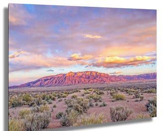 May include: A landscape photograph of a mountain range at sunset. The mountains are a reddish-brown colour, and the sky is filled with pink, orange, and blue clouds. The foreground features desert vegetation.