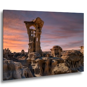 Puede incluir: Fotografía de un paisaje desértico con formaciones rocosas de arenisca únicas. La imagen presenta una paleta de colores cálidos de naranjas, rosas y marrones, sobre un cielo de atardecer. Las formaciones rocosas tienen formas y texturas únicas.