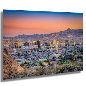 May include: A vibrant cityscape at sunset, featuring a mix of high-rise buildings and residential areas. The sky transitions from a deep blue to warm orange hues, with mountains in the background. The image captures a sense of urban life and natural beauty.