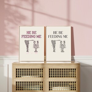 May include: Two framed prints with the text "He be feeding me" in pink and black lettering. Each print features a black and white illustration of a lobster holding a fork.