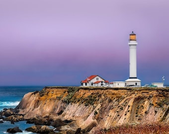 May include: A white lighthouse stands tall on a cliff overlooking a blue ocean with waves crashing against the rocky shore. The sky is a soft purple, creating a serene and picturesque scene.