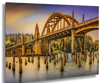 May include: A scenic photograph of a bridge over water. The bridge is made of concrete and has an arched design. The water is calm, and there are wooden posts in the water. The sky is a mix of orange, yellow, and blue.