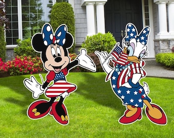 May include: Two cartoon characters, Minnie Mouse and Daisy Duck, dressed in red, white, and blue patriotic outfits. Minnie Mouse is wearing a red dress with white polka dots and a blue bow. Daisy Duck is wearing a blue dress with white stars and a red bow. Both characters are holding American flags.