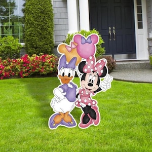 Może przedstawiać: Para kartonowych wycinanek przedstawiających Minnie Mouse i Daisy Duck. Minnie ma na sobie czerwoną sukienkę w białe kropki z różową kokardką i trzyma różowy balon. Daisy ma na sobie fioletową sukienkę z żółtą kokardką i trzyma żółty balon. Wycinanki stoją na zielonej trawie przed domem.