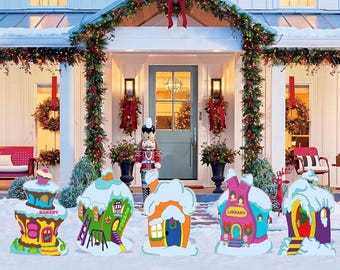 May include: A set of five colorful, flat, decorative yard signs featuring whimsical buildings including a bakery, school, house, library, and cottage, all topped with white snow, arranged in front of a house decorated for the holidays.