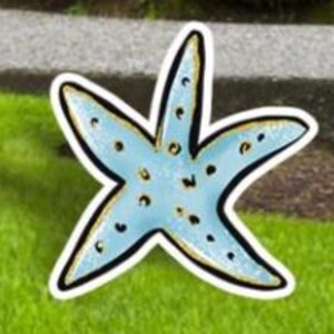 May include: A light blue starfish-shaped decoration with a black outline and gold accents. The starfish has small, dark blue spots and is placed on a green grassy surface.
