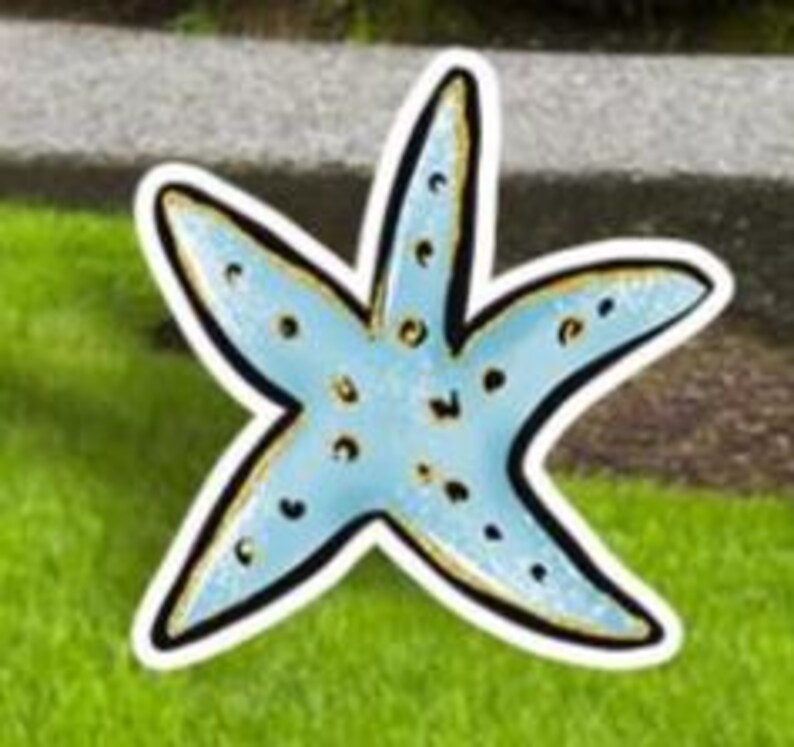 May include: A light blue starfish-shaped decoration with a black outline and gold accents. The starfish has small, dark blue spots and is placed on a green grassy surface.