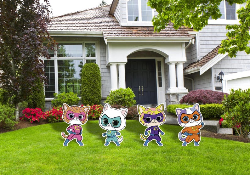 May include: Four colourful cat-shaped yard decorations stand on a green lawn in front of a house. Each cat has a superhero mask and costume in shades of pink, blue, yellow, and orange. The house has a grey exterior and a white porch.