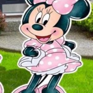 May include: A cutout of Minnie Mouse, a cartoon character, in a pink polka-dot dress and bow. The character has black ears, a smiling face, and is standing on a green lawn. The cutout is likely for outdoor decoration.