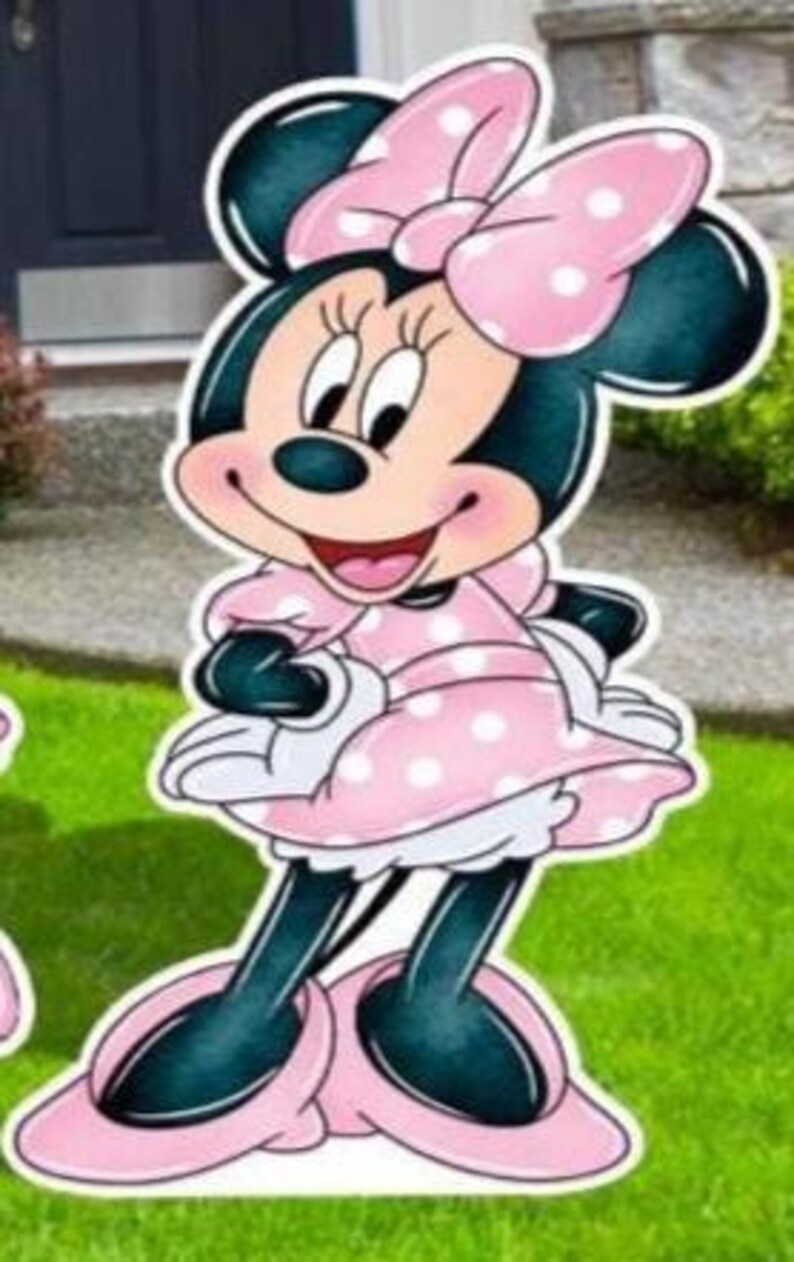 May include: A cutout of Minnie Mouse, a cartoon character, in a pink polka-dot dress and bow. The character has black ears, a smiling face, and is standing on a green lawn. The cutout is likely for outdoor decoration.