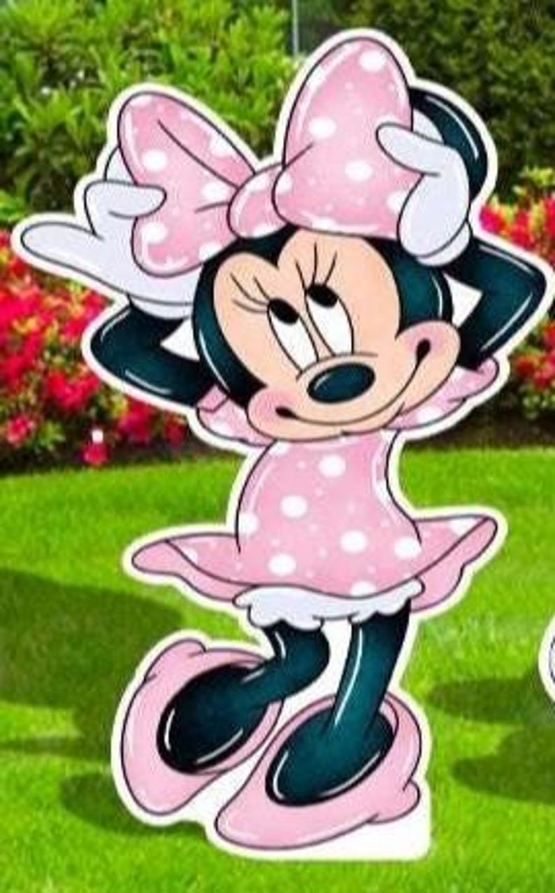 May include: A cutout of Minnie Mouse, featuring a pink polka-dot dress and bow. The character has black ears, a black nose, and white gloves. The cutout is standing on a green lawn with red flowers in the background.