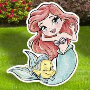 May include: A cartoon illustration of a mermaid with red hair, a seashell top, and a teal tail, accompanied by a yellow fish. The image is a cut-out, likely for yard or garden decoration, set against a green lawn and foliage.