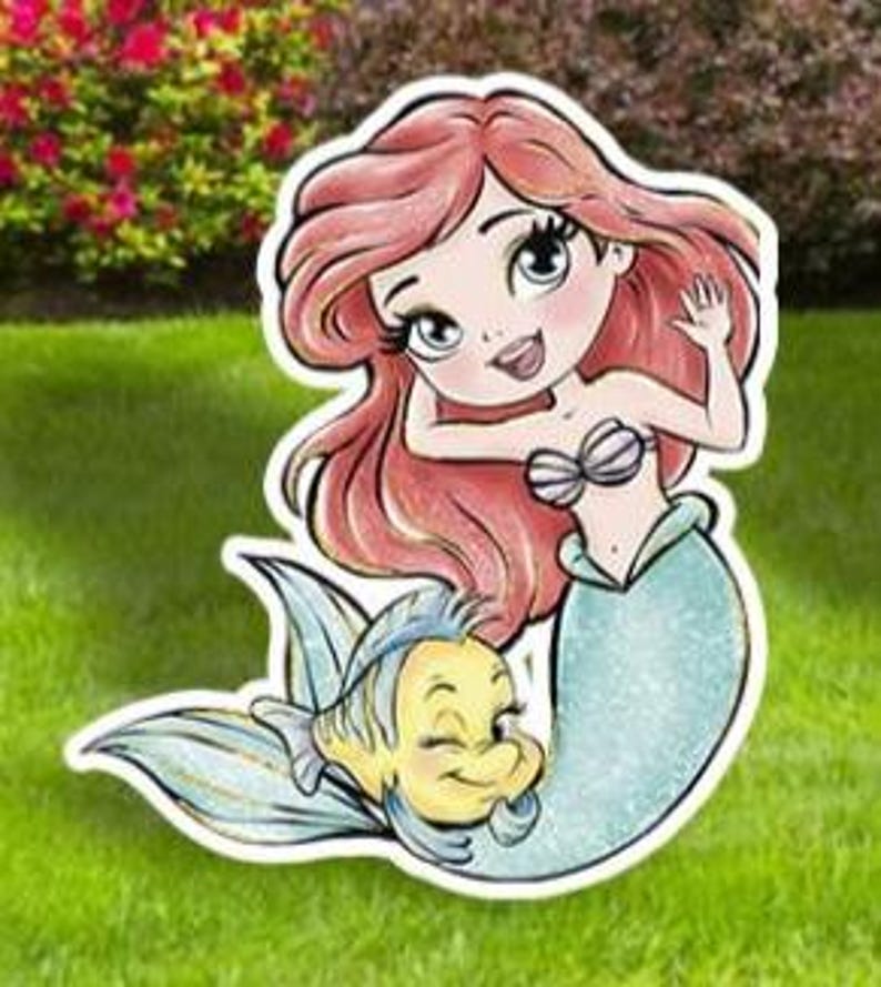 May include: A cartoon illustration of a mermaid with red hair, a seashell top, and a teal tail, accompanied by a yellow fish. The image is a cut-out, likely for yard or garden decoration, set against a green lawn and foliage.