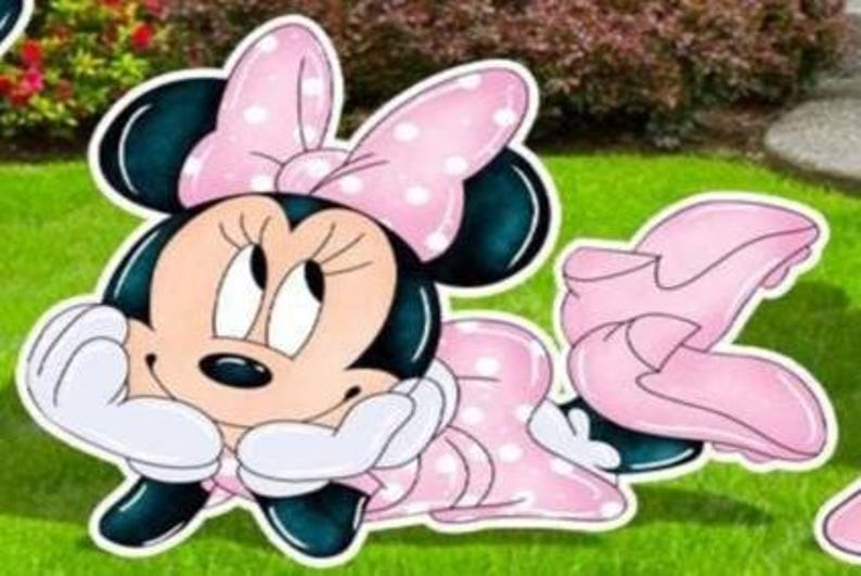 May include: A cartoon cutout of Minnie Mouse, featuring a pink and white polka-dot bow, dress, and shoes. Minnie Mouse is depicted with a tan face, black ears, and white gloves, resting her chin on her hands.