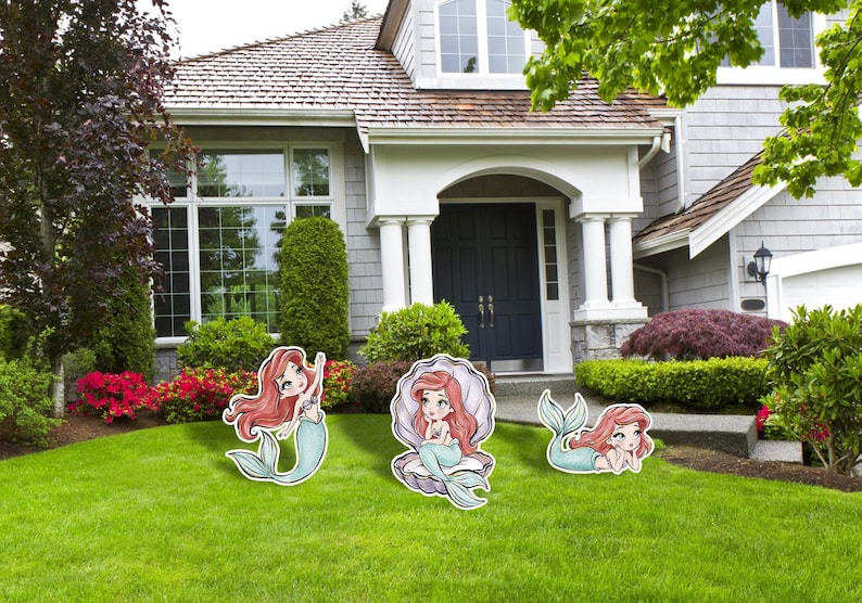 May include: Three mermaid yard decorations with red hair and teal tails are displayed on a green lawn in front of a house. One mermaid is standing, one is in a shell, and one is lying down.