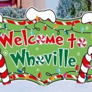 Może przedstawiać: Zielono-czerwony znak świąteczny z napisem "Witamy w Whoville" otoczony cukierkami w kształcie laski i światełkami.