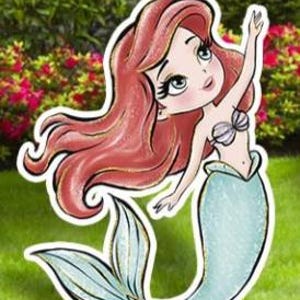 May include: A cartoon illustration of a mermaid with long red hair, a seashell bra, and a teal tail. The mermaid is waving with one arm raised. The image is a cutout, likely for outdoor decoration, set against a backdrop of green grass and bushes.