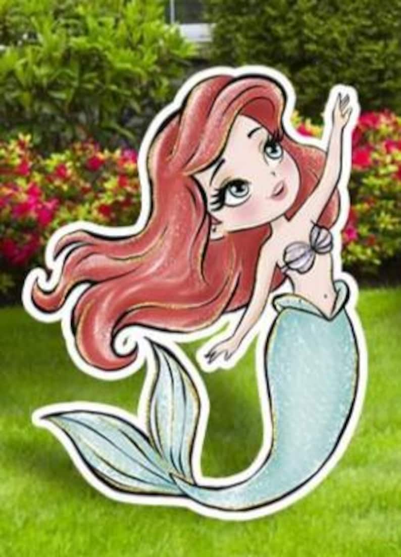 May include: A cartoon illustration of a mermaid with long red hair, a seashell bra, and a teal tail. The mermaid is waving with one arm raised. The image is a cutout, likely for outdoor decoration, set against a backdrop of green grass and bushes.