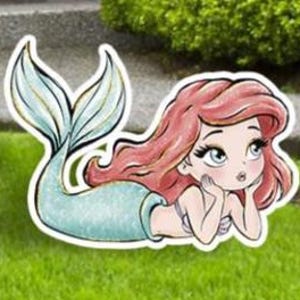 May include: A whimsical outdoor decoration featuring a cartoon mermaid with flowing red hair and a turquoise tail. The mermaid is lying down, resting her chin on her hands, and is set against a backdrop of green grass.