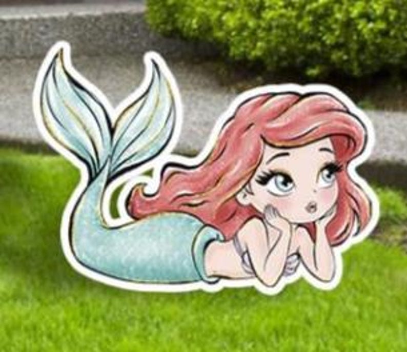May include: A whimsical outdoor decoration featuring a cartoon mermaid with flowing red hair and a turquoise tail. The mermaid is lying down, resting her chin on her hands, and is set against a backdrop of green grass.