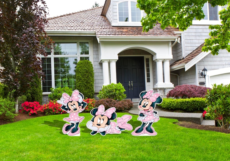 May include: Three Minnie Mouse yard decorations in various poses. Each figure is pink with black accents and a polka dot bow. They are placed on a green lawn in front of a house with a white exterior and a dark blue door.