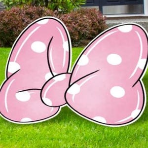 May include: A large, pink bow-shaped yard decoration with white polka dots. The bow has a black outline and is set on a green lawn. The bow is a whimsical outdoor decoration.