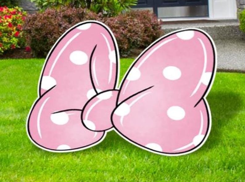 May include: A large, pink bow-shaped yard decoration with white polka dots. The bow has a black outline and is set on a green lawn. The bow is a whimsical outdoor decoration.