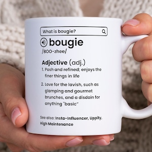 May include: White ceramic mug with a black definition of the word "bougie" printed on it. The definition includes the pronunciation, "BOO-zhee", and two definitions of the word. The mug is being held by a person's hand.