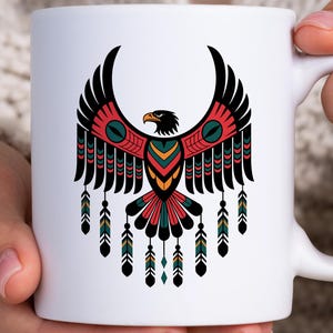 May include: White ceramic mug featuring a stylized eagle design in black, red, green, and yellow. The eagle has outstretched wings and decorative feathers. The mug has a white handle.