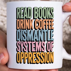 Puede incluir: Taza de cerámica blanca con el texto "READ BOOKS DRINK COFFEE DISMANTLE SYSTEMS OF OPPRESSION" en letras de bloque verdes, naranjas, azules, rosas y amarillas. La taza está sujeta por una persona.