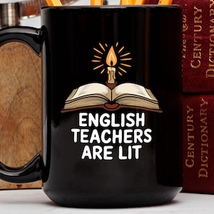 Puede incluir: Taza de café negra con un diseño blanco de un libro con una vela encendida en la parte superior. El texto en la taza dice "English Teachers Are Lit".