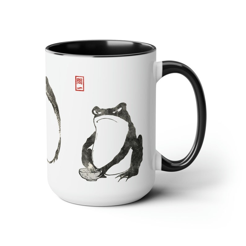 May include: White ceramic mug with a black rim and handle. The mug features a black and white ink illustration of a frog holding a bowl and chopsticks. The frog is sitting on a white background with a red square containing a Japanese character.