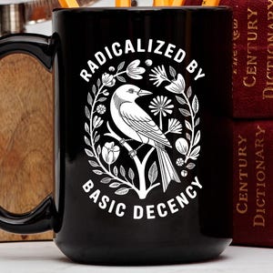May include: Black ceramic mug with a white graphic of a bird perched on a branch, surrounded by floral elements. The text "RADICALIZED BY BASIC DECENCY" is displayed in a circular arrangement. The mug has a large handle.