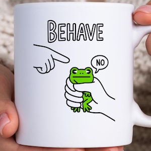 May include: White ceramic mug with the word "BEHAVE" in black block letters. A cartoon frog is held in a hand, with a speech bubble saying "NO". A finger points at the frog. The mug has a white handle.