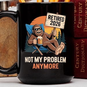 Op de afbeelding: Zwarte keramische mok met een cartoon Bigfoot die ontspant in een strandstoel, een biertje vasthoudt en een zonnebril draagt. De mok heeft de tekst "Retired 2026" en "Not My Problem Anymore."