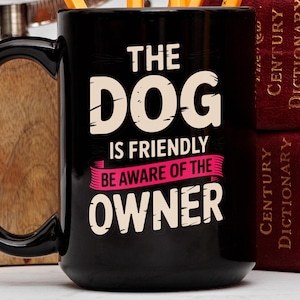 May include: Black ceramic mug with the text "THE DOG IS FRIENDLY BE AWARE OF THE OWNER" in white and pink. The mug has a large handle and is next to two red books.