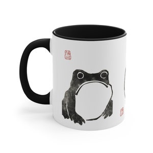 May include: White ceramic mug with a black handle and rim. The mug features a black and white illustration of a frog with a textured look. There are two red stamps on the mug.