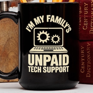 Puede incluir: Taza de café negra con el texto blanco "I'M MY FAMILY'S UNPAID TECH SUPPORT". El diseño incluye un gráfico de portátil con iconos de engranajes. La taza tiene un asa grande y está junto a una superficie de madera y libros.
