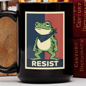Może przedstawiać: Czarny ceramiczny kubek z kreskówkową żabą w niebieskiej chuście i napisem "RESIST" poniżej. Żaba jest zielona z czarnymi kropkami. Kubek ma duży czarny uchwyt.