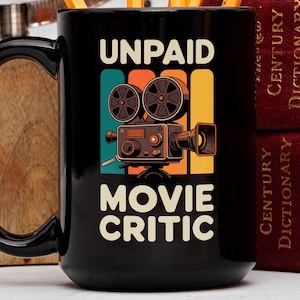 May include: Black ceramic mug featuring the text "UNPAID MOVIE CRITIC" with a vintage film camera graphic. The design includes teal, orange, and yellow accents. The mug has a large handle and is next to two burgundy books.