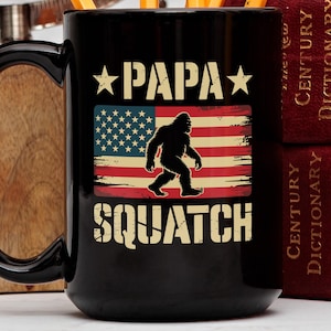 May include: Black ceramic mug featuring the text "PAPA SQUATCH" in white, with a Bigfoot silhouette over an American flag design. The mug has a large handle and is next to a stack of red books.